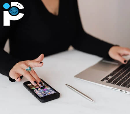 A lady looks at a smartphone on a desk which is next to a laptop